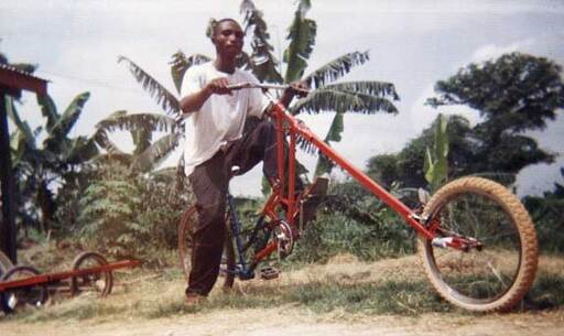 A man astride a modified orange bicycle in tropical Ghana, the front fork of the bike was elongated in a 'chopper' style, similar to a modified chopper motorcycle.